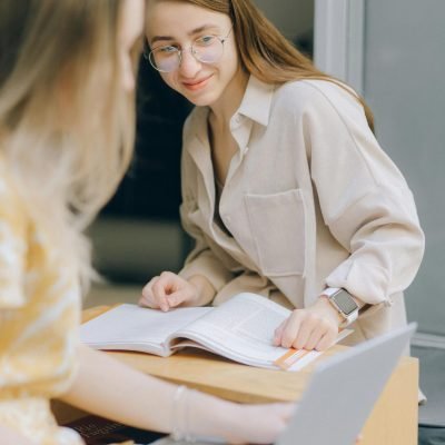 Two women sitting together, one using a laptop, the other looking at a book, in a collaborative workspace.