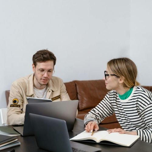 Two coworkers discussing a project with laptops and notes in a stylish office setting.