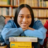 Smiling student in a library leaning on a stack of books, symbolizing education and learning.