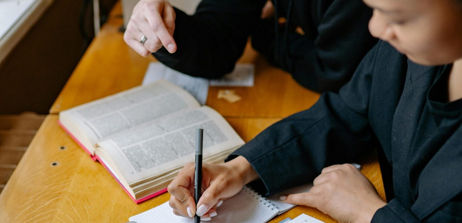 Two students studying together with books and a laptop, taking notes in a cozy indoor setting.